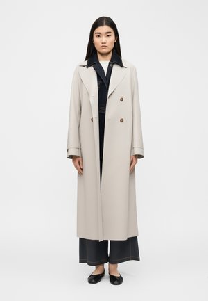 Femeie cu păr negru lung poartă un trench bej cu dublu rând de nasturi peste pantaloni evazați în culoare închisă și balerini negri, stând în fața unui fundal simplu.