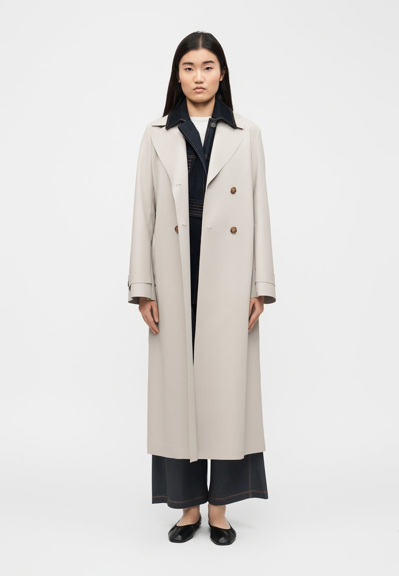 Γυναίκα με μακριά μαύρα μαλλιά φοράει ένα μπεζ διπλόμπουτο παλτό τύπου trench πάνω από σκούρο παντελόνι φαρδιάς γραμμής και μαύρα φλατ παπούτσια, στέκεται μπροστά σε απλό φόντο.