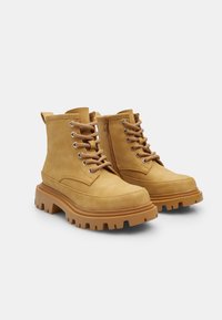 Bottines en suedine beige avec une épaisse semelle en caoutchouc, un laçage à l'avant et une fermeture éclair sur le côté. Présente une conception robuste et un bout arrondi.