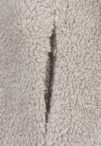 Lähikuva vaaleanbeigestä, pehmeästä ja kiharakarvaisesta kankaan pinnasta, jossa on pystysuuntainen sauma tai aukko keskellä.