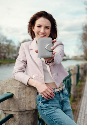 Gray leather wallet with a geometric zipper pull, featuring a smooth texture and embossed logo. Held prominently against a blurred landscape.