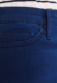 Mörkblå denimtyg med en slät yta, med en framficka, bältesöglor och en metallknappdetalj i midjan.