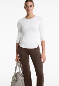Frau in einem taillierten weißen Langarmshirt und braunen Hosen, die in ihrer linken Hand eine große graue gesteppte Tasche hält.