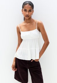 Hvid, letvægts ærmeløs bluse med en samlet bodice og delikat broderi ved kanten, parret med mørk bourgogne shorts.