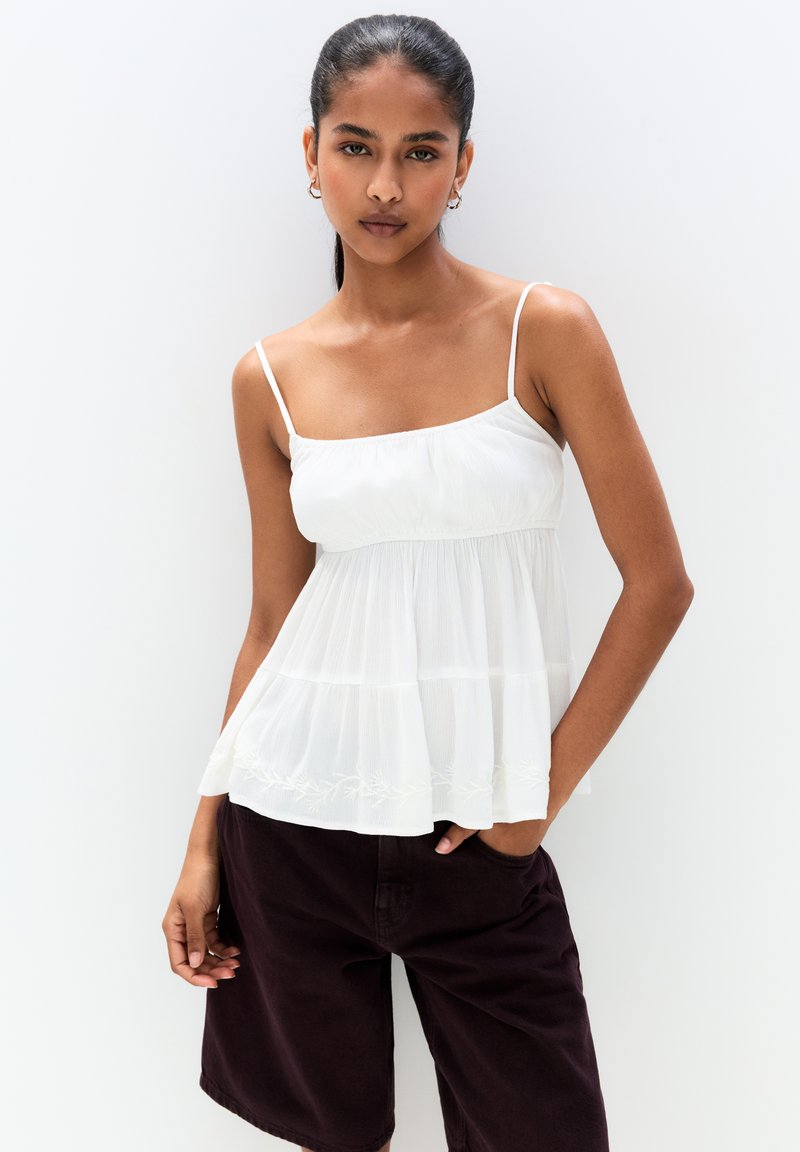 Hvid, letvægts ærmeløs bluse med en samlet bodice og delikat broderi ved kanten, parret med mørk bourgogne shorts.