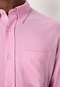 Vaaleanpunaisesta kankaasta valmistettu nappipaita, jossa on terävä kaulus, nappilista edessä ja rintatasku. Sileä pinta ja yksivärinen muotoilu.