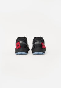 Zwarte sportieve schoenen met een gestructureerde bovenkant, reflecterende accenten en een rood logo-embleem op de hiel. Blauwe rubberen loopzool voor grip.