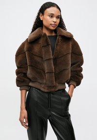 Brązowa kurtka z futerka sztucznego z szerokim kołnierzem, warstwową teksturą i prążkowanymi wykończeniami. Noszona na czarnych skórzanych spodniach, podkreślając skrócony fason.