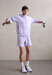 Lys lilla hettegenser med matchende shorts, holder vekter. Har en avslappet passform og ribbede mansjetter. Hvite sneakers og sokker.
