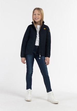 Marineblaue Fleecejacke mit hohem Kragen, Reißverschluss und kleinem gelben Regenschirm-Logo. Kombiniert mit dunkelblauen Jeans und weißen Sneakern.