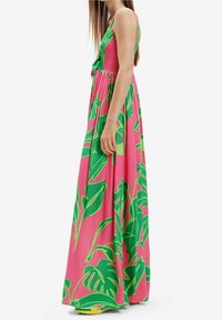 Robe maxi en tissu rose vif avec de grands imprimés de feuilles vertes, dotée de fines bretelles et d'une taille froncée pour une silhouette fluide.