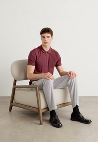 Bordová polo tričko, svetlosivé nohavice, čierne mokasíny, posedené na béžovom kresle s drevenými nohami, minimalistické pozadie.