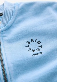 Sudadera azul claro con cuello acanalado y cremallera, que presenta un logotipo circular negro con el texto "LA SAINT" y "LONDON" en el frente. Textura suave.