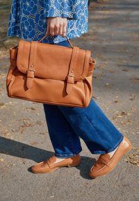 Brązowa skórzana torebka z dwoma klamrami i patką, zestawiona z jasnobrązowymi mokasynami. Niebieskie szerokie dżinsy z lekko postrzępionymi brzegami.