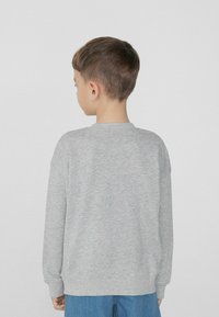 Šedá mikina s dlouhými rukávy, kulatým výstřihem a žebrovanými manžetami. Hladký materiál, volný střih, nošena s modrými džíny.