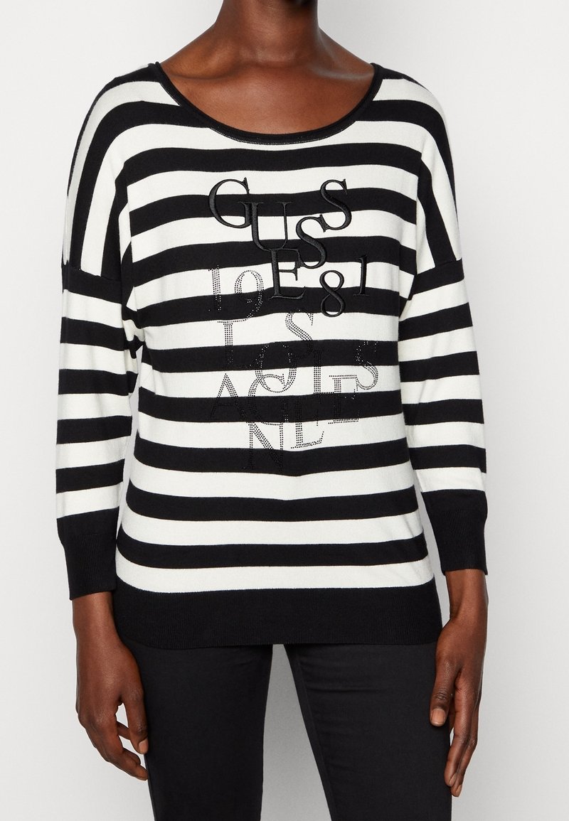 Pull à manches longues rayé noir et blanc avec le texte « GUESS 1981 LOS ANGELES », porté par une personne debout devant un fond uni.