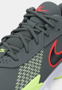 Grijze sport schoen met een bovenwerk van mesh, rode en zwarte accenten, gestructureerde overlays en een felgroene buitenzool. Bevat veters en merkdetails.