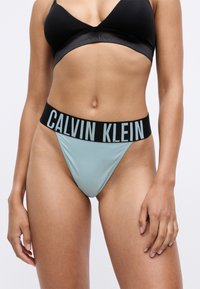 Calvin Klein bikinibroekjes van lichtblauwe stof met een zwarte elastische tailleband voorzien van witte letters. Minimalistisch ontwerp, zachte textuur.