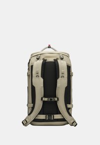 Sac à dos avec un extérieur en tissu beige et des accents en maille noire. Il dispose de bretelles et de boucles ajustables, avec une surface texturée pour une meilleure prise.