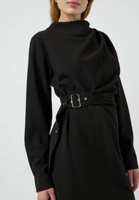 Černé šaty s dlouhými rukávy a vysokým výstřihem. Obsahují opasek s kovovou sponou a hladkou, matnou texturu.