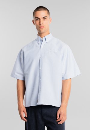 Muž s krátkymi vlasmi, oblečený v svetlomodrej a bielo pruhovanej košeli s krátkym rukávom na gombíky a tmavých nohaviciach, stojaci pred sivým pozadím.