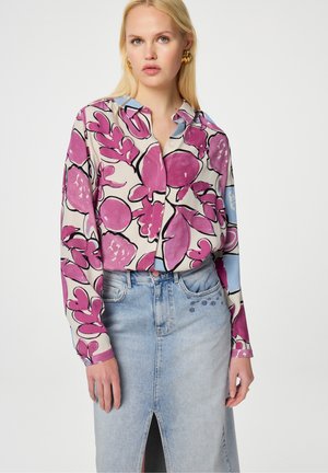 Bunt gemusterte Bluse mit pinkem Blumenmuster, Knopfverschluss vorne und hellblauem Kragen; kombiniert mit einem knielangen Jeansrock mit einem Schlitz vorne.