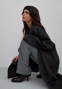 Zwarte laarzen met hoge hakken, grijze broek met wijde pijpen en een oversized donkerjasje. De outfit heeft zachte texturen en vloeiende lijnen.