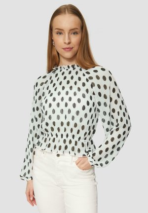 Blouse blanche transparente avec des pois noirs, col froncé et taille peplum. Présente des manches longues avec des poignets élastiques, portée avec un pantalon blanc.