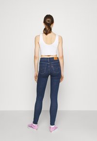 Calças de ganga skinny azul escuro com cintura alta, apresentando um patch de couro no verso e costuras subtis nos bolsos. Combinadas com ténis cor-de-rosa.