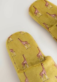 Yellow slippers featuring a textured, soft fabric, adorned with a repeated giraffe pattern. Rounded toe and open back design.