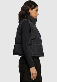Svart puffervest med høy krage, frontglidelås og lilla detaljer. Har en løsere passform og kviltet tekstur, kombinert med en svart topp.