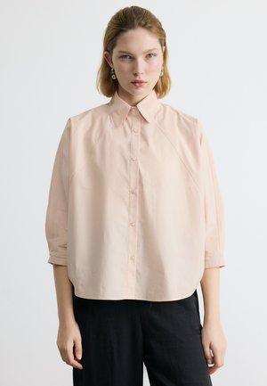 Ženska s kratkimi blond lasmi, ki nosi srebrne uhane, ohlapno bledo roza srajco z gumbi in črne hlače na enostavni beli podlagi.