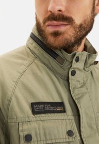 Olive green jacket with a high collar, featuring a zip and snap closures, and a black patch reading "MADE FOR DAILY ADVENTURES."