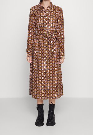 Femme portant une robe à manches longues, mi-mollet, à motifs géométriques en marron, blanc et violet, associée à des bottes noires à lacets.