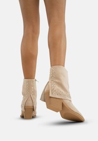 Botas de tobillo de ante beige con punta afilada, decoradas con tachuelas metálicas a lo largo de las costuras y un tacón cuadrado medio.