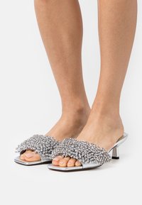 Sandales à talons argentées avec une tige texturée ornée de petites perles décoratives. Design à bout carré et un petit talon aiguille.