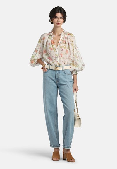 Žena u prozirnoj cvjetnoj bluzi s nabranim rukavima, svijetlo plavim trapericama, smeđim sandalama na petu i koja drži bijelu torbicu sa zlatnim lancem.