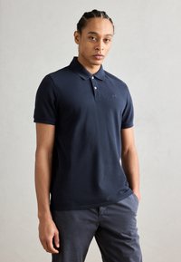 Jeune homme avec des cheveux tressés, portant un polo bleu marine à manches courtes et un pantalon gris foncé, se tenant avec une main dans la poche.
