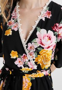 Mustapukuinen kietaisumekko, jossa on värikkäitä kukkakuoseja vaaleanpunaisena ja keltaisena. Syvä V-pääntie ja pitsireunus pääntiellä. Sidottu vyötärölle.