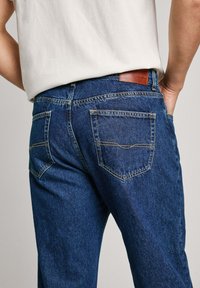 Jeans de mezclilla azul oscuro con corte recto, que presentan dos bolsillos traseros y costuras en contraste. Logo de parche de cuero en la cintura.