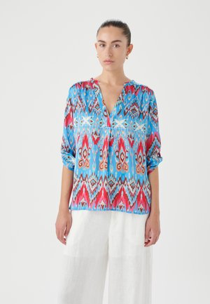 Blusa leggera blu con motivi geometrici rossi e marroni, scollo a V, maniche arrotolabili e vestibilità comoda. Abbinata a pantaloni in lino bianchi.