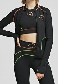 Černý atletický outfit skládající se z texturované zipové bundy a odpovídajícího crop topu. Mezi detaily se objevují neonově zelené akcenty a loga značky v oranžové barvě.