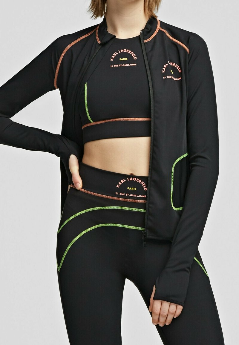 Černý atletický outfit skládající se z texturované zipové bundy a odpovídajícího crop topu. Mezi detaily se objevují neonově zelené akcenty a loga značky v oranžové barvě.