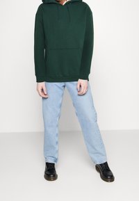 Felpa verde scuro con tasca frontale, abbinata a jeans a gamba dritta blu chiaro e stivali neri stringati. La felpa presenta un cappuccio regolabile con cordino.