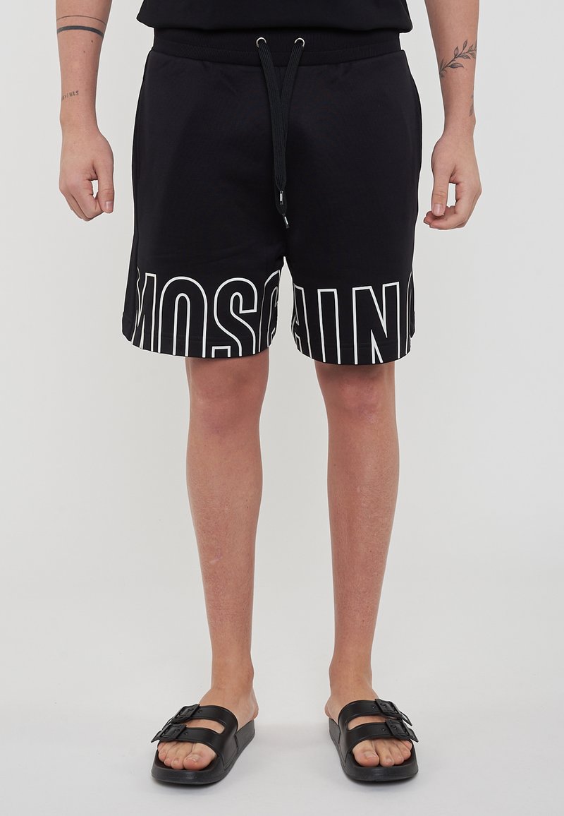 Pantaloni corti neri con vita elasticizzata e logo "MOSCHINO" bianco stampato sull'orlo. Texture liscia, taglio fino al ginocchio, abbinati a sandali neri.