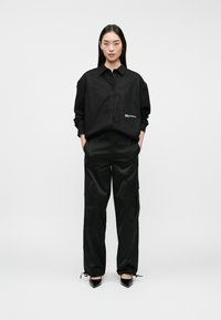 Camisa negra de botones con detalle de logo, combinada con pantalones cargo negros que cuentan con bolsillos, tobillos ajustados y tacones negros brillantes.