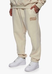 Kremfargede joggebukser i myk tekstil, med sidelommer og en subtil trykt logo. Kombinert med hvite joggesko.