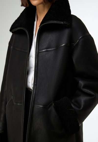 Veste en cuir noire avec un grand col en shearling noir, fermeture éclair visible et poches latérales ; présente une texture lisse et un design structuré.