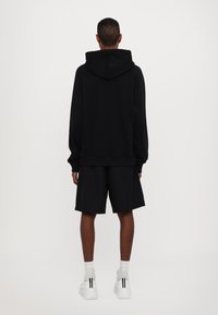 Μαύρη φούτερ μπλούζα με κουκούλα και χαλαρή εφαρμογή, σε συνδυασμό με μαύρες βερμούδες. Χαρακτηριστικά περιλαμβάνουν απαλή υφή και λευκά αθλητικά παπούτσια με μαύρες λεπτομέρειες.