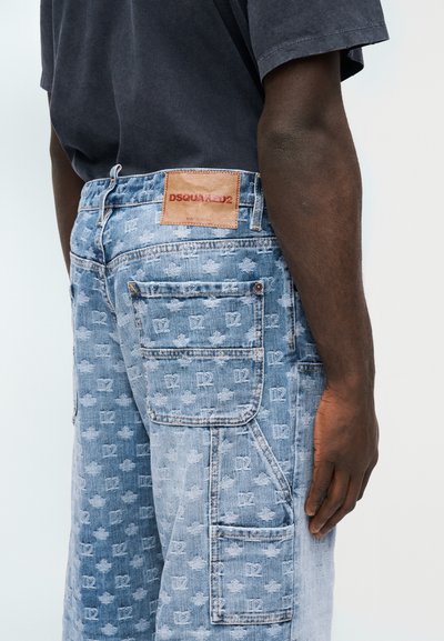 Άτομο που φοράει μπλε τζιν cargo με επαναλαμβανόμενα μοτίβα "D2" και φύλλο σφενδάμου, καθώς και δερμάτινη ετικέτα DSQUARED2 στη ζώνη μέσης.